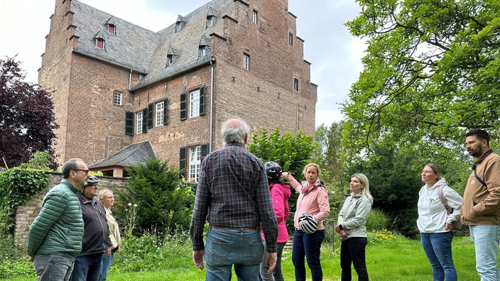 Mehrere Menschen stehen in einem Kreis vor einem Schloss in einem Garten. 