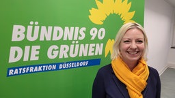 Einen Plakat der Grünen, davor eine lächelnde Frau mit blonden Haaren, dunklem Blazer und orangefarbenem Schal