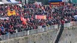 Eine Menschenmenge mit Bannern und Plakaten stehen auf einem Platz nahe dem Rhein in Köln