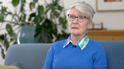 Eine ältere Frau mit Brille sitzt auf einer Couch und trägt einen blauen Pullover.