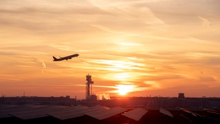 Zu sehen ist ein Flugzeug, welches in der Dämmerung über Häuser fliegt.