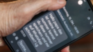 Ein Finger tippt auf ein Smartphone-Display. Darauf ist eine Betrugs-SMS zu lesen. Inhalt der SMS: "Hallo Papa, das ist meine neue Nummer, kannst Du diese Nummer speichern und mir auf Whatsapp schreiben? LG"