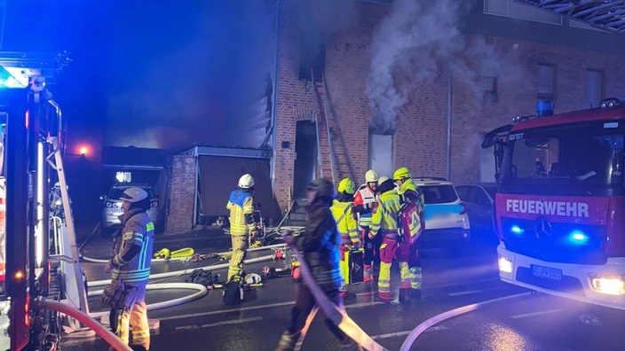 Einsatzkräfte und Fahrzeuge der Feuerwehr stehen vor dem brennenden Wohnhaus in Eschweiler. Aus Fenstern kommt Rauch. An eines ist eine Leiter angelehnt.