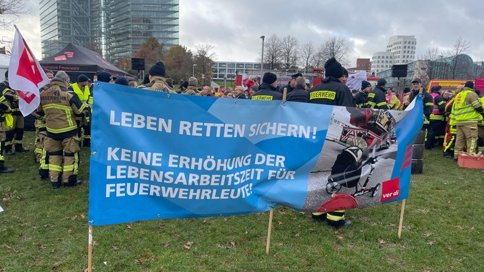 Die Feuerwehrleute bei ihrer Mahnwache auf der Wiese vor dem Landtag. Auf einem zentralen Plakat steht: "Leben retten sichern! Keine Erhöhung der Lebensarbeitszeit für Feuerwehrleute!"