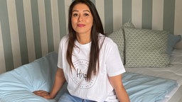 Eine junge Frau mit braunen Haaren und in weißem T-Shirt sitzt auf einem Bett vor einer gestreiften Wand und lächelt