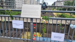 Ein an einem Geländer montierte Gedenktafel, daneben Blumen und weiße Blätter mit Zitaten
