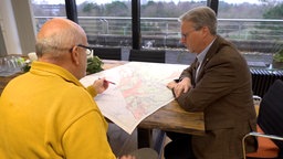 Zwei Männer sitzen an einem Tisch und schauen auf einen Stadtplan.