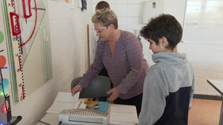 Eine Frau mit blonder Kurzhaarfrisur steht neben einem Kind mit dunklen Haaren an einem Tisch und zeigt ihm etwas