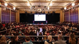 Blick in einen vollen Veranstaltungsaal mit Bühne und Leinwand 