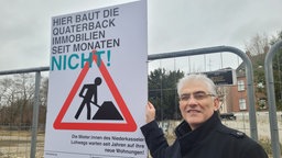 Ein Mann mit weißen Haaren und Brille steht in einem Bauzaun mit großem montierten Schild