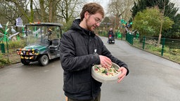 Ein Mitarbeiter des Zoos steht auf dem Gehweg, in der Hand eine Futterschüssel. Im Hintergrund steht ein Fahrzeug.