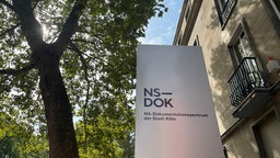 Schild mit dem Schriftzug "NS-DOK - NS-Dokumentationszentrum der Stadt Köln" hängt an der Außenfassade des NS-Dokumentationszentrums.