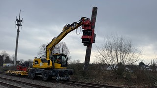 Ein gelber Bagger fährt auf Schienen, in seinem Greifarm hält er ein sechs Meter langes Stahlrohr.