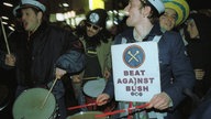 Verkleidete Männer mit Trommeln, verschiedenen Hüten und einem Schild "Beat against Bush"