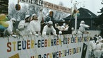 Viele Männer in weißen Kutten auf einem Karnevalswagen, auf dem bunte Buchstaben stehen