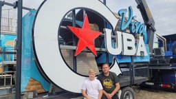 Ein Mann und sein Sohn sitzen vor einem großen Schild "Viva Cuba"