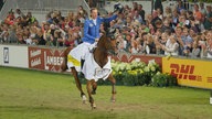 Ein Mann reitet auf einem braunen Pferd durch ein Stadion. Die Zuschauer winken ihm zu.