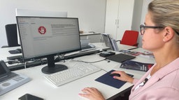 Eine Frau mit rosa Blauer und brille sitzt an einem Computer. Auf dem Bildschirm ist ein Stoppschild-Ähnliches Icon, in dem ZeMAG steht