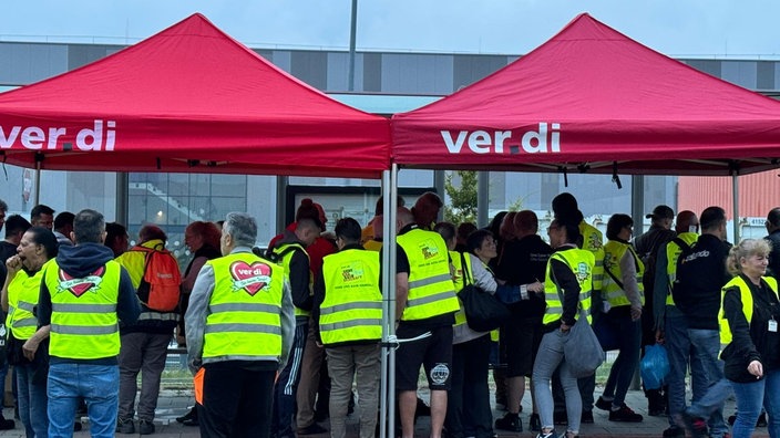 Viele Mitarbeiter mit gelben Warnwesten auf dem Bürgersteig unter zwei roten Pavillons