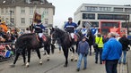 Auf dem Foto sind Menschen in blau-schwarzen Jacken, die auf schwarzen Pferden sitzen und den Anfang des Karnevalszuges bilden.