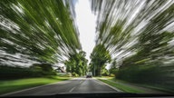 Eine Straße wird aus einem fahrenden Auto heraus gesehen, am Rand sind Bäume verschwommen zu erkennen.