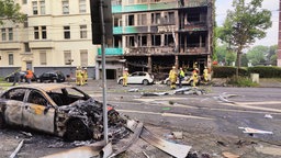 Feuerwehrleute stehen vor dem ausgebranntem Mehrfamilienhaus. Ein ebenfalls ausgebranntes Auto ist im Vordergrund zu sehen.