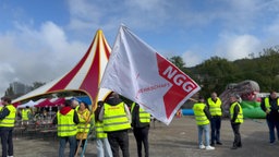 Im Fordergrund weht eine Fahne der Gewerkschaft NGG, im Hintergrund sind Streikende in gelben Warnwesten zu sehen