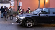 Ein großes schwarzes Auto steht vor der Schule. Davor stehen mehrere Kinder und zwei Schülerlotsen in gelben Westen.