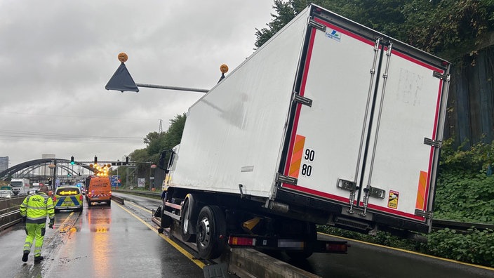 Ein Laster steht auf einer Leitplanke. Um den LKW stehen Rettungsfahrzeuge. Man sieht die Rückansicht.