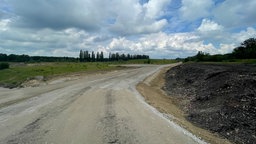 Das Brachgelände, ein Schotterweg und hinten ein paar Bäume