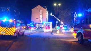 Mehrer Fahrzeuge der Feuerwehr stehen vor einem Haus. Eine Drehleiter wird an dieses haus rangefahren. 