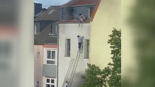 Ein Haus aus dem Rauch aufsteigt. Eine Person reicht ein Kind von einem Balkon, zu einer weiteren Person auf einer Leiter herunter.