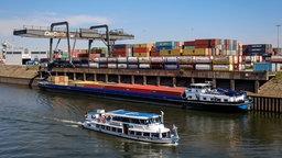 Auf dem Foto ist eine Fähre auf einem Kanal im Vordergrund. Dahinter ist ein Frachtschiff, das über einen Kran mit Containern beladen wird.