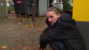 Die Obdachlose sitzt neben einem Kassenautomaten auf einem Parkplatz