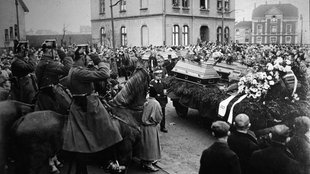 Ein Schwarz-Weiß-Foto zeigt mehrere Pferdewagen, auf denen Särge stehen, gesäumt von einer Menschenmenge. Ein Reiter in Uniform salutiert. 