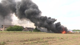 Auf dem Foto sind Gebäude hinter einem Feld, die brennen und in einer riesigen schwarzen Rauchwolke verschwinden.