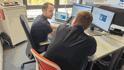 Die Mitarbeiter des Dezernats für Zentrale Dienste und Digitalisierung sitzen an einem Schreibtisch vor 2 Computer-/ und einem Laptopbildschirm und schauen sich an.