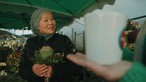 Eine Blumenverkäuferin hält einen Blumenstrauß in der Hand. Im Vordergrund ist die Lokalzeit Tasse zu erkennen.