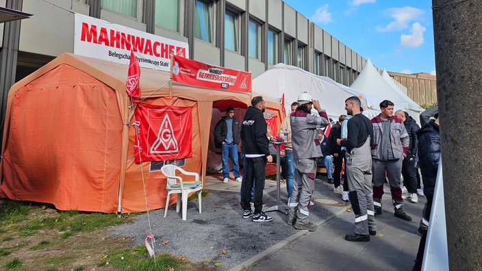 Ein orangenes Zelt an dem Banner der IG Metalol befestigt sind. Vor dem Zelt halten sich mehrer Personen auf.