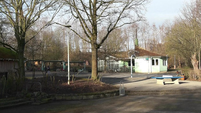 Ein Schulhof mit Bäumen, einer Tischtennisplatte, einem Fahrradständer und einem kleinen grünen Gebäude im Hintergrund.