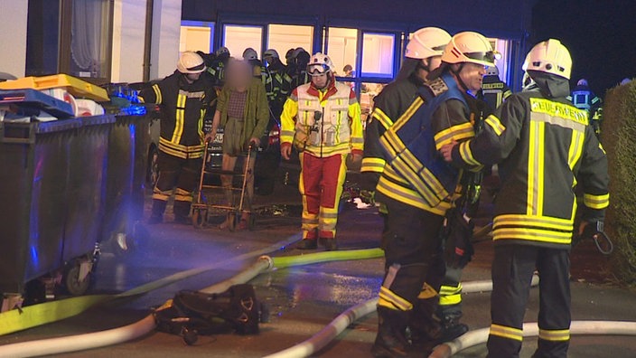 Auf dem Foto sind etliche Feuerwehrleute vor einem Seniorenheim. Zwei führen einen Mann mit Rollator ab.