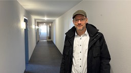 Zu sehen ist Andreas Hohlfeld im Flur des ehemaligen Hotels. Er trägt ein weißes Hemd, eine schwarze Jacke und eine graue Mütze.