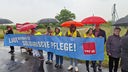Zahlreiche Menschen stehen an einer Autobahnauffahrt. Sie halten Regenschirme, sowie ein Plakat mit der Aufschrift "Laut für solidarische Pflege!" hoch.