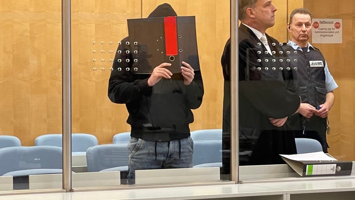 Der Angeklagte steht im Gerichtssaal und hält sich einen Aktenordner vor das Gesicht, neben ihm steht sein Verteidiger