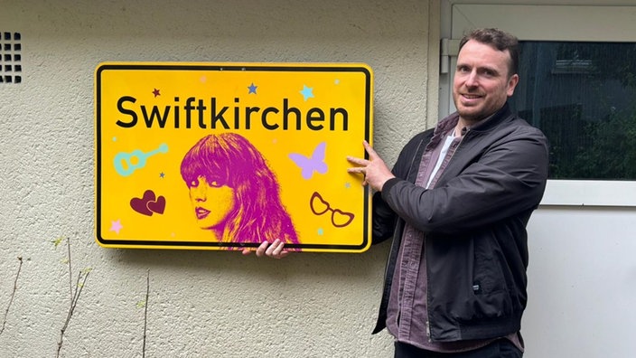 Ein Mann präsentiert ein gelbes "Swiftkirchen"-Schild. Er lacht.