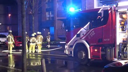 Ein Feuerwehrauto mit Blaulicht steht vor einem Mehrfamilienhaus, daneben mehrere Feuerwehrleute