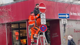 Zwei Arbeiter stehen auf einer Leiter und montieren von einem Mast ein Schilder ab