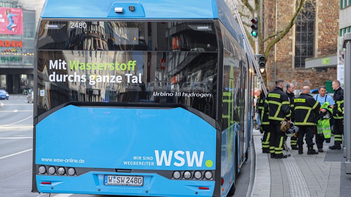 Ein blauer Linienbus von hinten. Darauf steht "Mit Wasserstoff durchs ganze Tal". Neben dem Bus stehen Feuerwehrleute