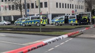 Auf dem Foto ist Absperrband zu sehen, dahinter mehrere Polizeiautos und Polizist:innen