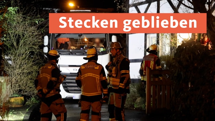 Feuerwehrleute stehen vor einem Sattelschlepper, der in einer Einfahrt stecken geblieben ist; ein rotes Banner mit weißer Schrift "Stecken geblieben" liegt auf dem Bild